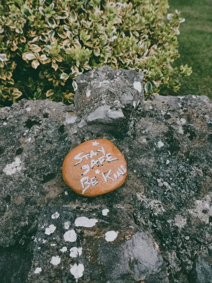 Pomalowany kamień z napisem „Stay Safe Be Kind” leżący na skale, na tle zieleni ogrodu